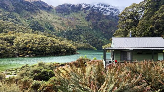 A small green hut with a chimney stands beside Lake Mackenzie on the Routeburn Track, New Zealand, alpine ferns in the foreground and a snow dusted bluff behind. Wide shot