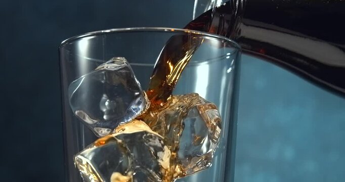 Cola pouring into glass with ice cubes in slow motion on blue background