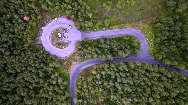 Cars navigating winding asphalt roads through dense alpine forest, parking at a circular trailhead with vibrant greenery and natural mountain scenery.