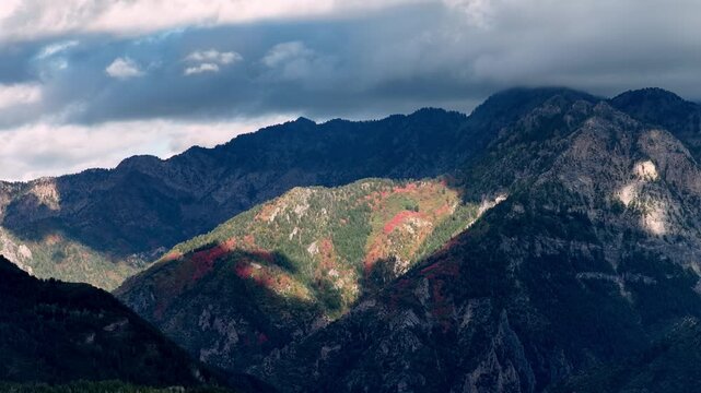 Clouds moving over a rugged alpine forest with vivid autumn colors and shifting shadows dancing across the steep mountainous terrain in a static time lapse.