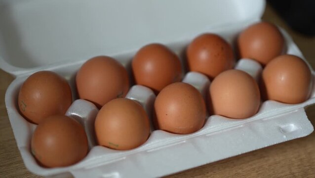 A close-up shot of ten brown chicken eggs arranged neatly in two rows inside an open white styrofoam carton box on a wooden surface.