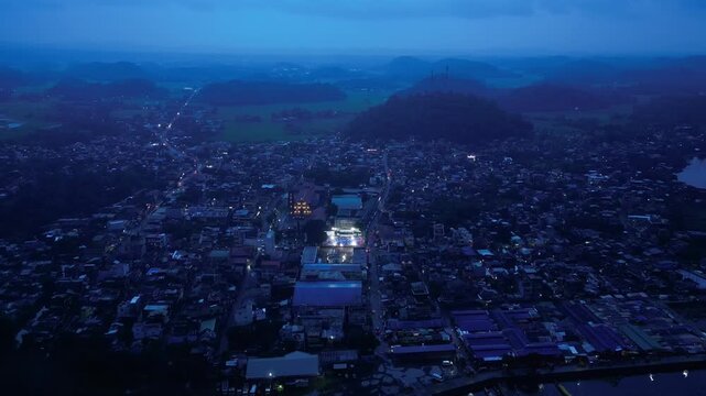 Cinematic drone view of an illuminated basketball court during a community event in Buhi, Camarines Sur, Philippines. Seen from far above and across the town at blue hour, the intense court lighting c