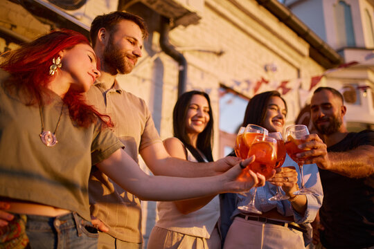 Five friends clink Aperol Spritz cocktails on rooftop terrace with festive joy. Friendship, social celebration and summer lifestyle concept.