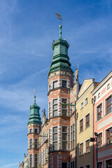 Great Armoury, a historic building featuring prominent green-domed towers and red brick facades, standing alongside colorful merchant houses on the vibrant Long Market street in Gdańsk, Poland © photoopus