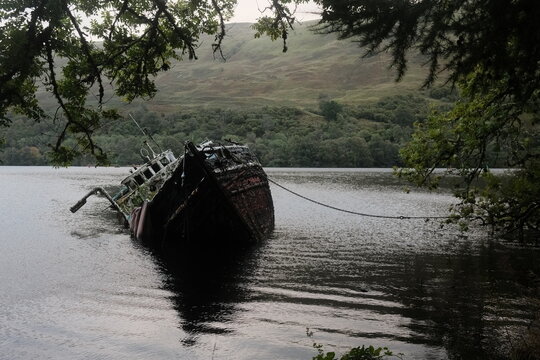 View of a partially submerged shipwreck in the calm waters of Loch Lochy, framed by overhanging tree branches and rolling green hills in the background, Blairgowrie, Scotland, United Kingdom.