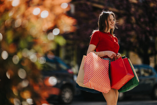 A smiling young woman carries multiple colorful shopping bags on a sunny city street. She walks past blurred cars and autumn foliage while holding patterned and plain paper bags.