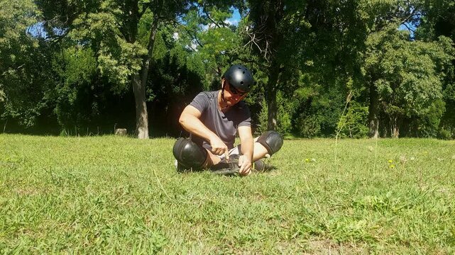 Full shot of a young man sitting on green grass in a city park removing and detaching a wheel from inline skates  during a break 