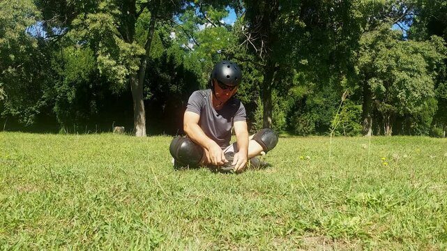 Full shot of a young man sitting on green grass in a city park installing and screwing a wheel back onto inline skates using a hex key during a break 