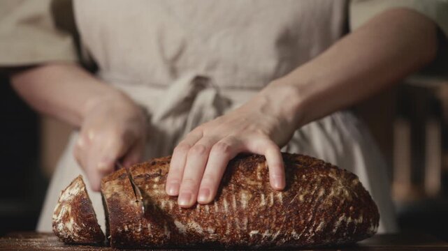 Close-up of baker in rustic linen apron slicing freshly baked artisan sourdough bread with knife on wooden cutting board. Homemade crusty loaf for breakfast, bakery promos, and healthy food concepts.