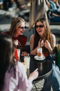 Two young women wearing sunglasses laugh and chat while sitting at an outdoor cafe table. The friends enjoy iced coffee and a relaxed conversation in a sunny urban setting.