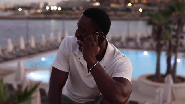 Man talking on smartphone at waterfront resort at dusk. Man talking on a smartphone in a waterfront setting at dusk, with a pool and city lights in the background.