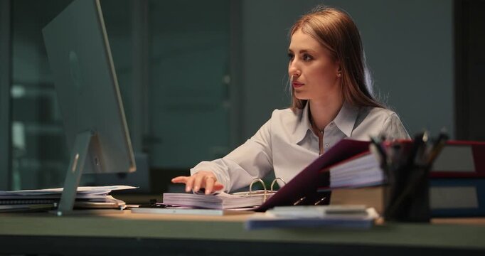 Focused office woman sits at busy desk with laptop and folders. Employee reviews paperwork continuing working alone during late evening shift