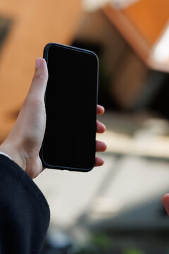 Hand holding a modern smartphone with a blank black screen outdoors, offering a template for app interfaces, digital content, or communication technology concepts