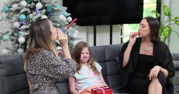 Young women sit with girl near decorated Christmas tree at home. Family group blows party horns preparing for New Year celebration with playful energy