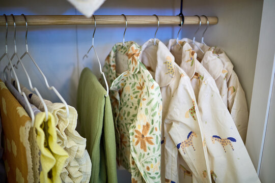 Assorted children's clothing in various colors and patterns including floral, gingham, and stripes hanging on racks and in closets, featuring cotton, linen and muslin fabrics in boutique or home setti