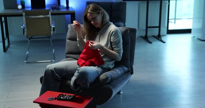 Woman sits in armchair with red fabric and sewing supplies nearby. Focused person stitches material with hand carefully working on clothing repair