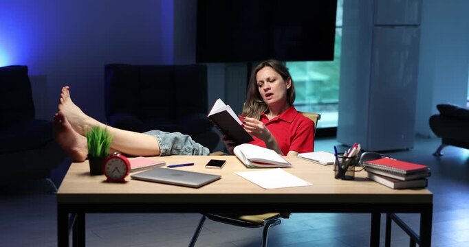 Tired office woman rests feet on desk with book in hand. Worker puts book aside stretching arms before scrolling through messages on smartphone