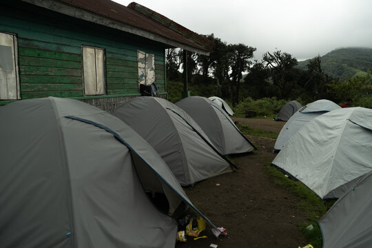 View of grey camping tents set up on a dirt field next to a rustic green wooden building with misty mountains in the background in Kohima, Nagaland, India.