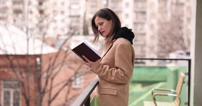 Woman in beige coat reads thick book while standing on balcony in autumn. Focused reader follows pages enjoying quiet reading outside in fresh air