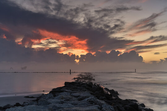 View of the rocky breakwater extending into the calm sea at sunrise with dramatic clouds and a vibrant orange sky at Lido di Dante, Emilia-Romagna, Italy.