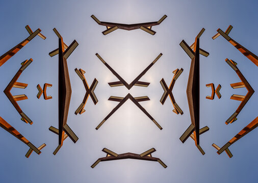 a symmetrical, kaleidoscope-style pattern made from architectural elements or wooden beams against a light blue sky