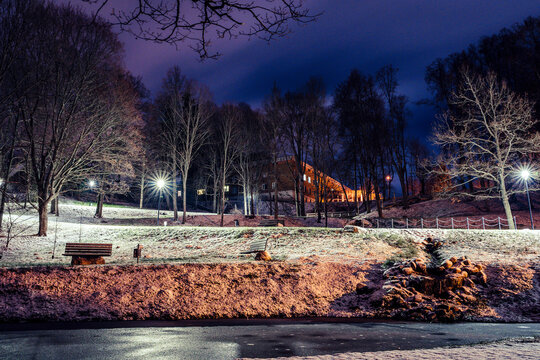 View of a park at night with benches, bare trees, and street lights illuminating a light dusting of snow on the ground, with a building visible in the background in Latvia.