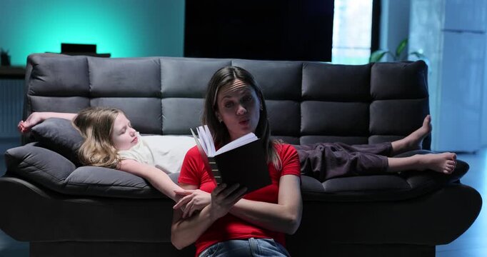 Mother reads bed time story to little girl sleeping on sofa slow motion. Caring woman with book tells fairy tale to preteen child in living room