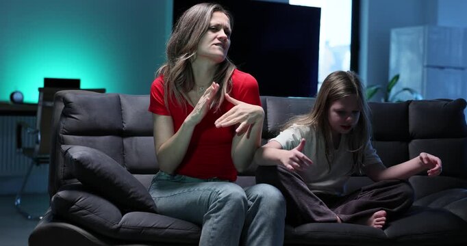 Happy mother and daughter dance grimaving as sit nearby on sofa slow motion. Family of woman and girl vibe to rap music together in living room