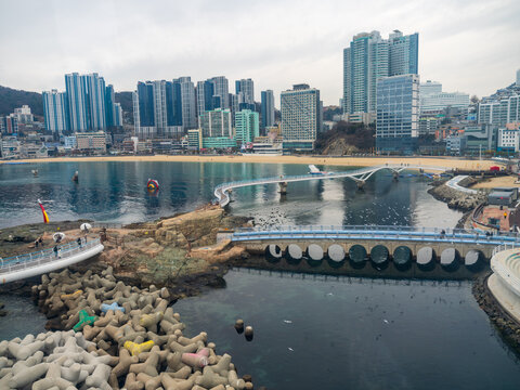 View of the Songdo Cloud Trails bridge extending over the sea with the modern skyline of Busan in the background, featuring concrete tetrapods and calm water in Busan, Busan, South Korea.
