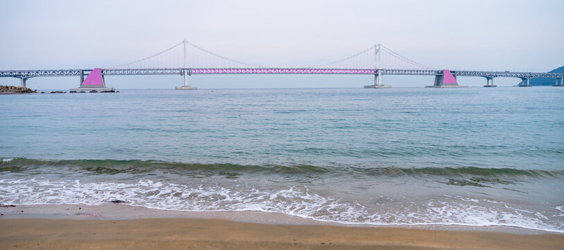 View of Gwangan Bridge spanning across the sea with calm waves washing onto the sandy beach under a soft overcast sky in Busan, Busan, South Korea.
