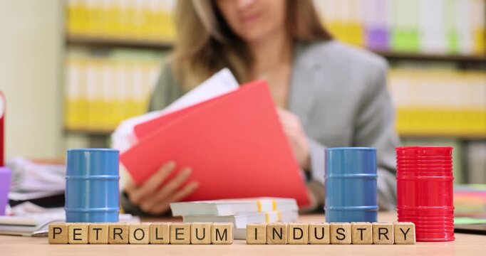 Phrase Petroleum Industry spelled on wooden cubes and miniature fuel barrels on desk. Female manager checks documents for oil trading in office