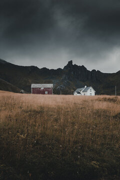 View of a red barn and a white house situated in a dry grassy field beneath dramatic dark mountains and a stormy sky in Napp, Nordland, Norway.