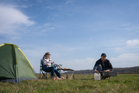 Couple relaxing outdoors on a sunny day during a camping trip, with one person lounging in a chair watching the other prepare meat on a portable grill beside their green tent