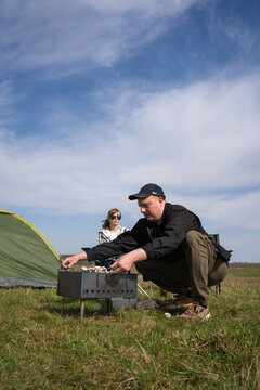 Happy couple spending leisure time together in a field, grilling food on a barbecue, sitting on camping chairs, enjoying a relaxing meal outdoors under a blue sky. Vertical photo