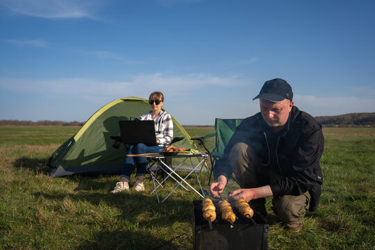 Couple camping in a field, woman working on laptop next to a tent while man prepares food on a portable barbecue grill under a clear blue sky, embracing digital nomad lifestyle and outdoor leisure