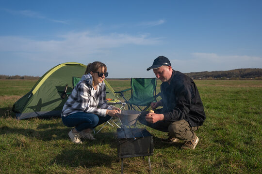 Couple preparing food on a small grill next to their tent in an open field, enjoying the freedom and simple lifestyle of an outdoor adventure and recreational activity