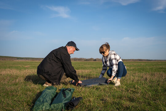 Adult couple setting up a tent together in a wide green meadow under a clear blue sky, preparing for a daytime camping adventure and enjoying outdoor teamwork and nature
