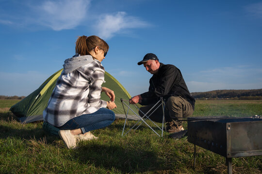 Couple collaborating to assemble a portable camping table in a grassy field under a blue sky, enjoying leisure and recreation during their outdoor adventure