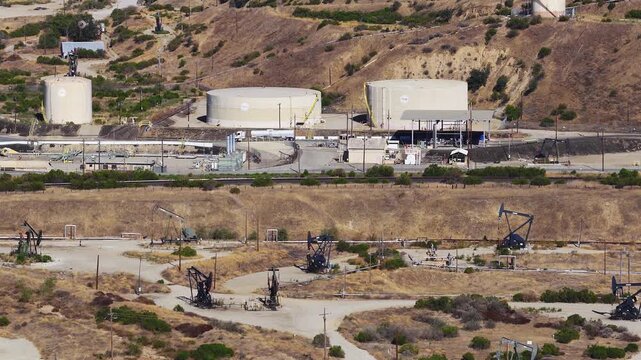 Aerial view shows pump jacks pumping crude petroleum alongside large storage tanks and industrial facilities across an active California oil field landscape.
