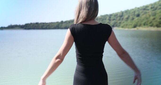 Woman in sportswear opens wide curtains of cozy lakeside cabin. Female steps onto wooden bridge enjoying breathtaking view of calm lake in morning