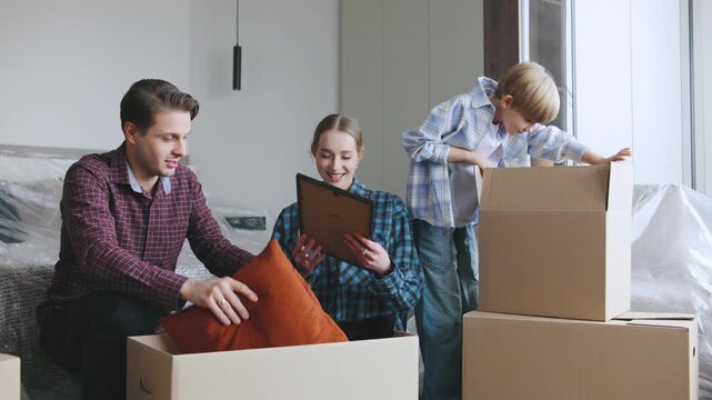 A family and their real estate agent unpack cardboard boxes in their new home. A couple discusses items while their son explores a box.