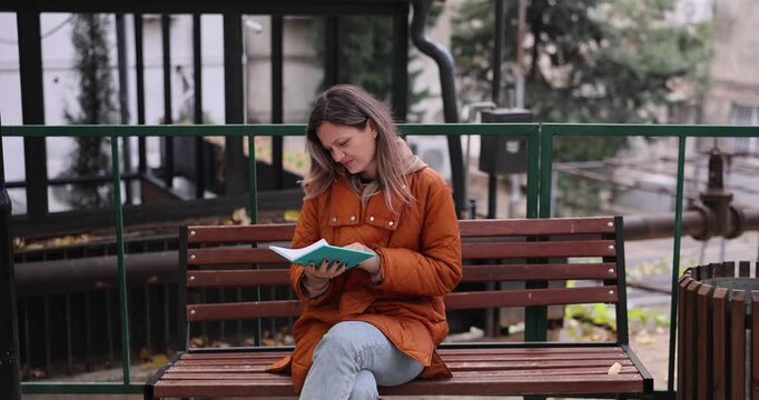 Relaxed reader completely absorbed in captivating small book. Stylish woman sits on city bench and carefully follows intriguing story. Slow motion