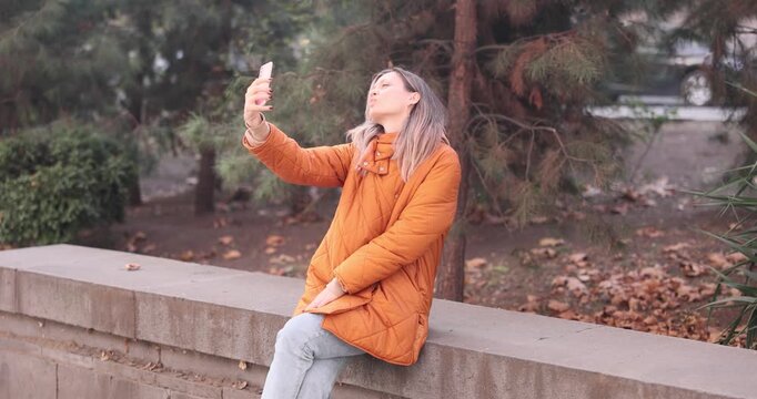 Joyful woman holds modern smartphone and takes selfie on parapet. Cheerful female in stylish autumn outfit smiles warmly at phone camera. Slow motion