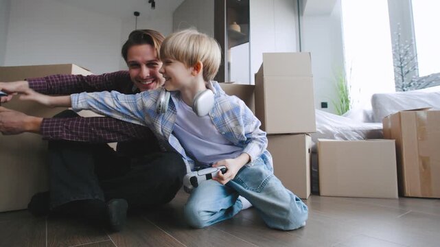 A man and his son sit on the floor and play video games in their new house. Cardboard boxes are piled around them as they settle in together.