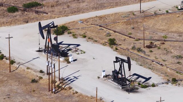 Aerial high view shows mechanical pump jacks actively extracting petroleum in an arid California oilfield with utility poles and pipes under direct sun.