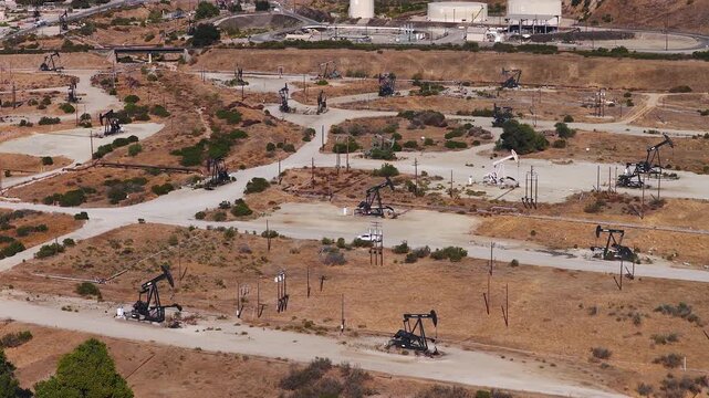 Aerial view captures numerous nodding donkey pump jacks actively extracting petroleum across an arid oil field landscape in California, with storage tanks and dirt roads visible.