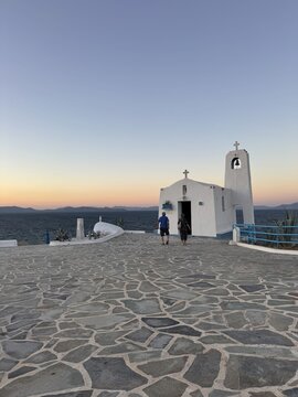Porto Rafti, Greece - 21 September 2026: View of a small white Greek chapel by the sea at sunset, with two people walking towards the entrance on a stone-paved courtyard.
