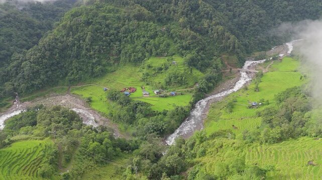 Drone shot of a scattered mountain hamlet beside a winding river surrounded by rice terraces and mist covered forest hills in Nepal