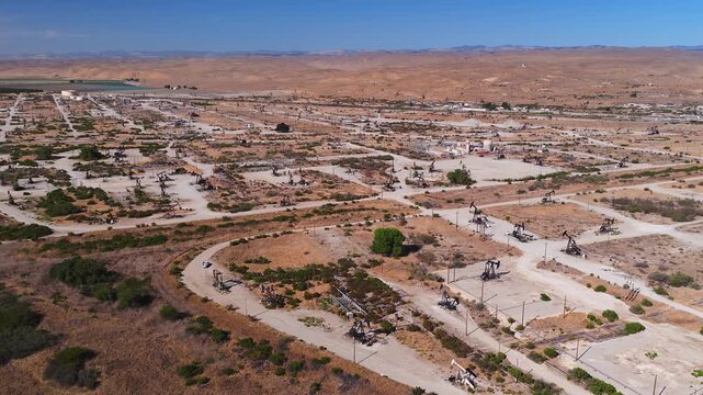 Aerial view over working pump jacks and industrial infrastructure across a vast arid oil field in California. The drone shot shows petroleum extraction equipment. Oil tariffs, trade war, price plunge.