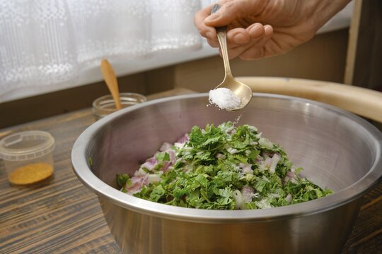 A hand adds salt to a bowl filled with minced meat, chopped onions and fresh herbs. The kitchen is bright, with sunlight streaming through the window, creating a warm atmosphere for cooking